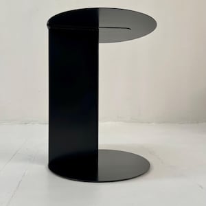 Könnte beinhalten: Ein moderner, minimalistischer Beistelltisch in mattschwarz. Der Tisch hat eine kreisförmige Basis, eine rechteckige vertikale Stütze und eine halbkreisförmige Tischplatte. Das Design ist schlicht und modern, passend für verschiedene Einrichtungsstile. Der Tisch ist ca. 46 cm hoch.