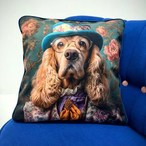 Può includere: Cuscino quadrato decorativo con un cane che indossa un cappello blu, occhiali rotondi e una giacca a fantasia floreale. Il cuscino ha uno sfondo verde scuro con fiori rosa e un bordo blu. Il pelo del cane è marrone chiaro.