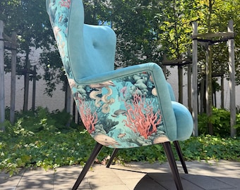 May include: A high-back wing chair featuring solid light blue upholstery on the front and a vibrant underwater-themed fabric on the back. The exterior pattern displays coral reefs, sea life, and fish in shades of pink, green, and blue. Four dark legs.