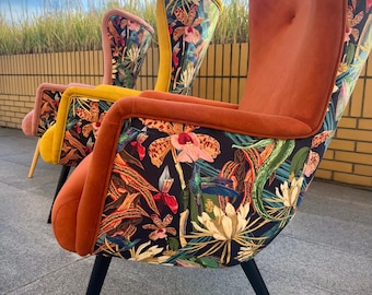 May include: Three wingback chairs with black tapered legs. The front chair is upholstered in burnt orange velvet with a black fabric back featuring a colorful botanical print of orchids, hummingbirds, and tropical leaves. Two similar chairs follow.