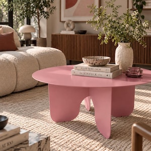 Op de afbeelding: Een ronde, roze salontafel met een glad oppervlak en vier gebogen, golfachtige poten. Op de tafel liggen twee gestapelde boeken met de titels A Life More Calm en The Kinfolk Home, een gespikkelde kom, een getextureerde witte vaas en een klein paars glazen schaaltje.