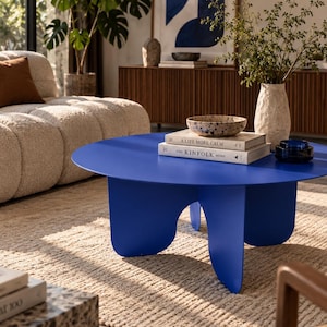 Könnte beinhalten: Ein runder, leuchtend blauer Couchtisch mit glatter Oberfläche und drei breiten, geschwungenen Beinen. Auf dem Tisch stehen eine gesprenkelte Schale, eine kleine dunkelblaue Glasschale und zwei Bücher mit den Titeln A Life More Calm und The Kinfolk Home.