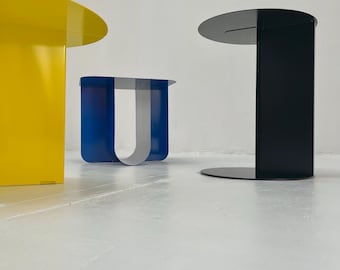 Op de afbeelding: Drie moderne bijzettafels in primaire kleuren. Een gele tafel met een rond blad, een blauwe tafel met een U-vormig ontwerp en een zwarte tafel met een rond blad en voet. De tafels zijn gemaakt van metaal en hebben een minimalistisch ontwerp.