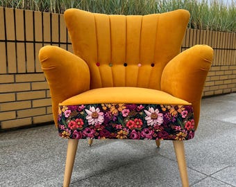 May include: A mustard yellow velvet armchair with a channel-tufted backrest featuring five small multicolored buttons. The front panel of the seat is upholstered in a dark fabric with a vibrant floral pattern. It stands on four light wood legs.