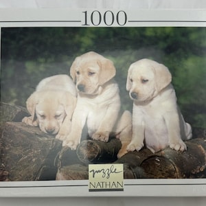 May include: A 1000-piece jigsaw puzzle featuring three adorable Labrador Retriever puppies. The puppies are light yellow and are sitting on a pile of logs. The puzzle is by Nathan.