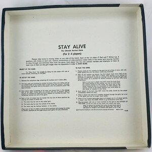 Pode incluir: Um jogo de tabuleiro vintage intitulado "STAY ALIVE" para 2-4 jogadores. O jogo inclui um tabuleiro com 14 slides, bolinhas de gude e bot&otilde;es. O objetivo &eacute; ser o &uacute;ltimo jogador com bolinhas de gude restantes na grade. A caixa do jogo &eacute; branca com texto preto.