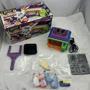 May include: A Creepy Crawlers Bug Maker toy set including a purple and green electric oven, a cardboard box with product graphics, a purple plastic tool, a black tray, four metal insect molds, and several small bottles of colored liquid plastic.