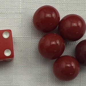 May include: A red die with white dots showing the number four, next to five red spheres. The objects are on a light gray textured surface. The spheres are glossy and reflect light.