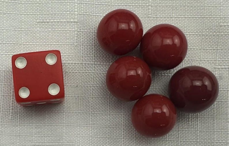 May include: A red die with white dots showing the number four, next to five red spheres. The objects are on a light gray textured surface. The spheres are glossy and reflect light.