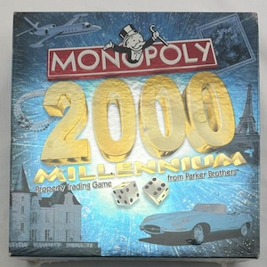 May include: Monopoly 2000 Millennium edition board game box. The box features the Monopoly logo, the number 2000 in gold, and illustrations of a jet, dice, a car, and the Eiffel Tower. The words "Property Trading Game" are also visible.