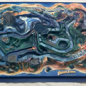 May include: Fireball Island board game. The game board is a detailed, three-dimensional island with a volcano, waterways, and pathways. The island is surrounded by a blue border. The game's title is printed on the bottom of the board.
