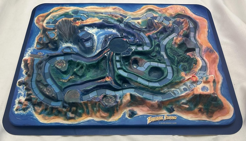 May include: Fireball Island board game. The game board is a detailed, three-dimensional island with a volcano, waterways, and pathways. The island is surrounded by a blue border. The game's title is printed on the bottom of the board.