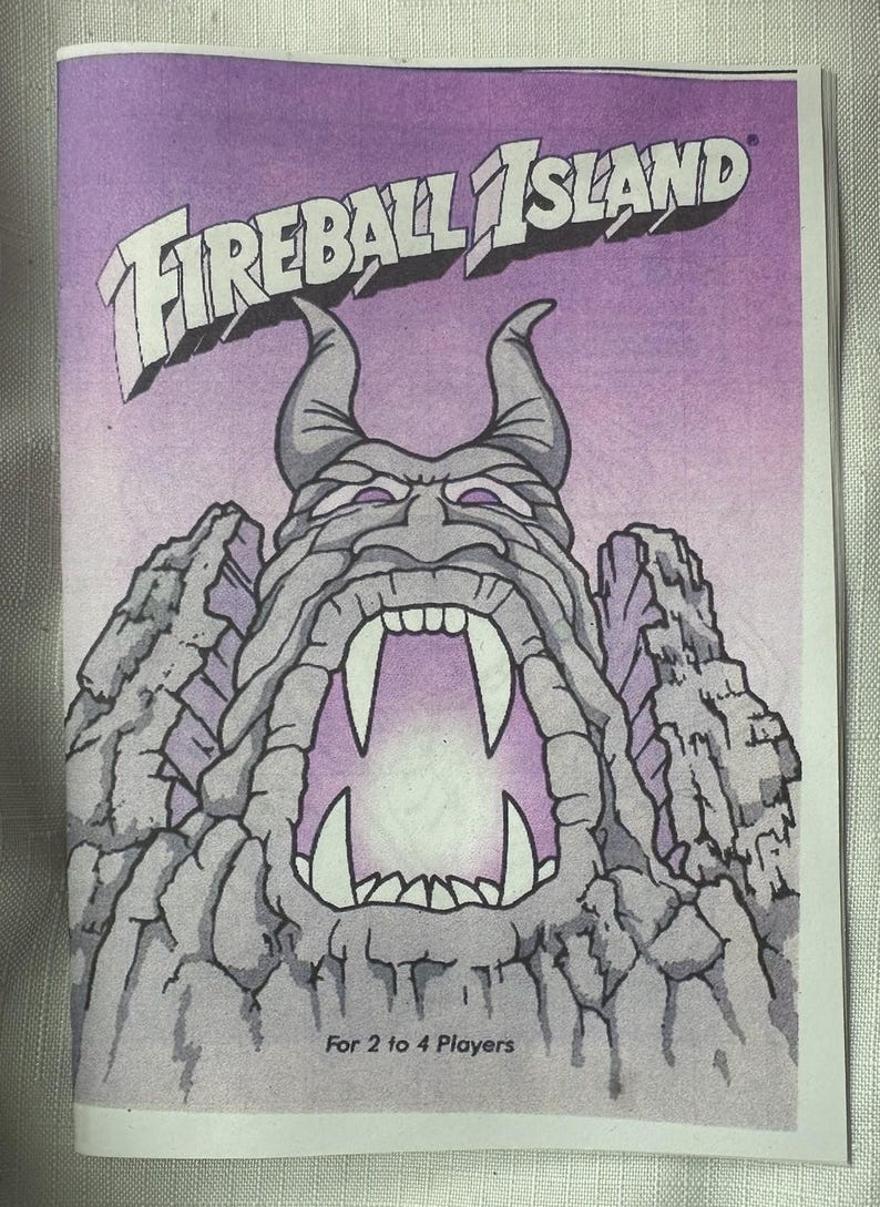 May include: Cover art for the board game Fireball Island. The image features a stylized illustration of a volcanic island with a menacing face, large fangs, and the game's title in bold letters. The text "For 2 to 4 Players" is also visible.