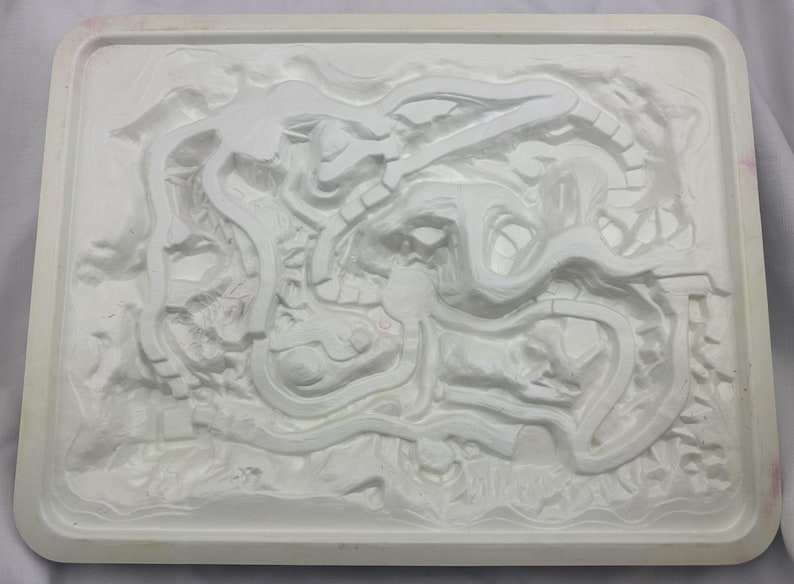 May include: A white, rectangular mold with a detailed, raised relief design. The design features winding, organic shapes and textures, possibly for casting or molding. The mold has a raised border.