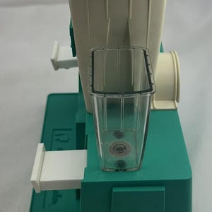 May include: A vintage food mill, featuring a cream-coloured hopper and a clear plastic collection cup. The base is teal green, and the mill has a white handle and a cylindrical attachment. It appears designed for processing fruits or vegetables.
