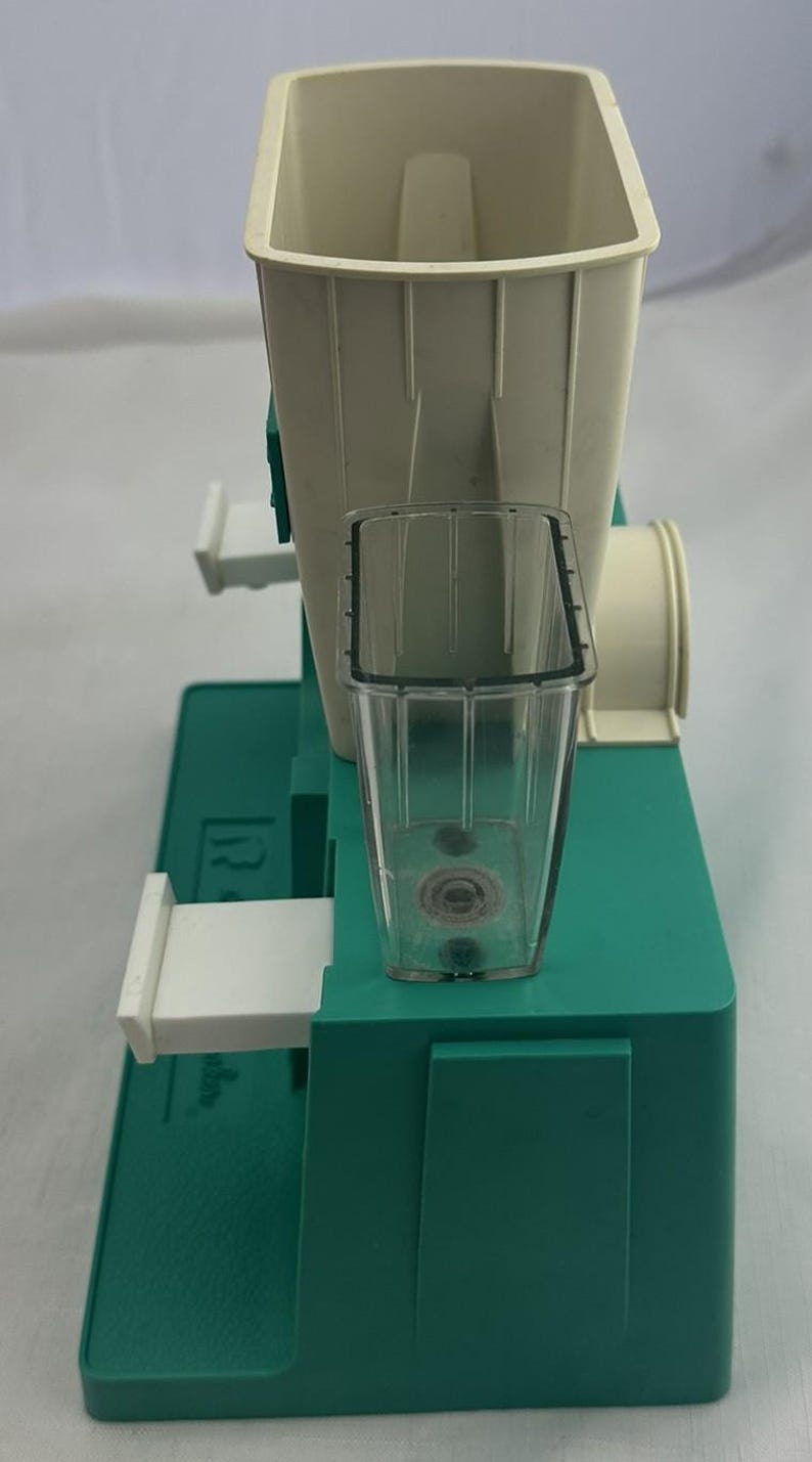 May include: A vintage food mill, featuring a cream-coloured hopper and a clear plastic collection cup. The base is teal green, and the mill has a white handle and a cylindrical attachment. It appears designed for processing fruits or vegetables.