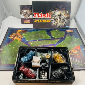 May include: Risk Star Wars Clone Wars Edition board game featuring a game box, a game board with a galactic map, a rulebook, and a plastic tray containing bags of blue, yellow, red, and black game pieces, dice, and a deck of cards.