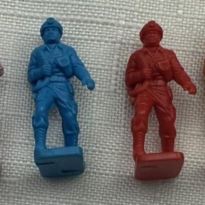 May include: Four toy soldier figurines in shades of purple, blue, red, and orange. Each soldier is standing on a rectangular base and wearing a helmet and uniform. The figures are made of plastic.
