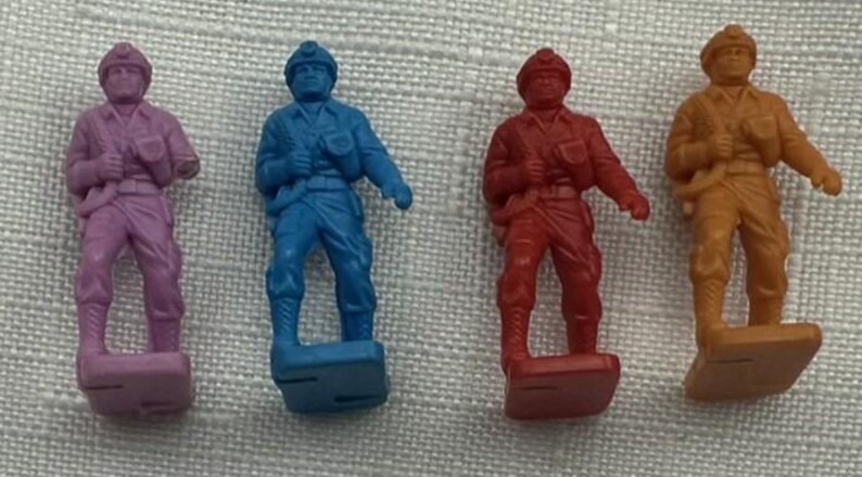 May include: Four toy soldier figurines in shades of purple, blue, red, and orange. Each soldier is standing on a rectangular base and wearing a helmet and uniform. The figures are made of plastic.