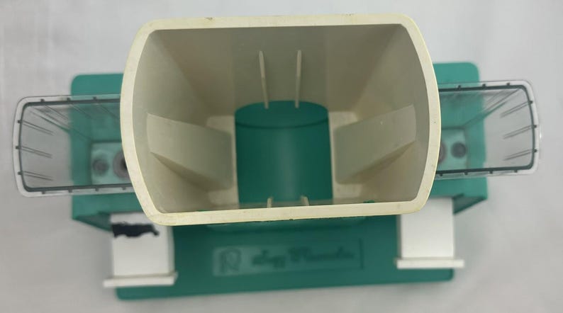 May include: An overhead view of a seed planter. The planter is primarily green and off-white, with a large, open hopper and two transparent seed compartments on either side. The base has the text "Slogg Products".