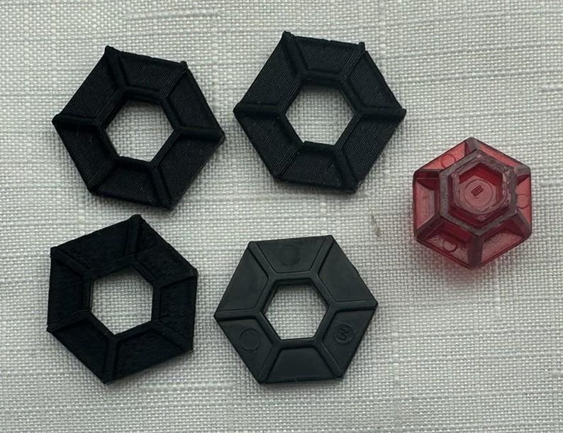 May include: An assortment of black and red hexagonal objects. Four black objects have a hollow center, while the red object is translucent. The objects are arranged on a textured, light gray surface.