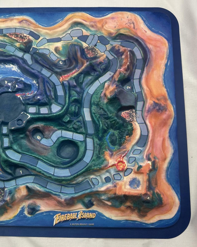 May include: A board game titled "Fireball Island" with a detailed island map design. The game board features a blue border, with a colorful island landscape in shades of green, brown, and orange. The game's title is displayed in white text.