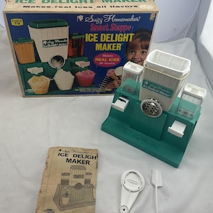 May include: A vintage Suzy Homemaker Sweet Shoppe Ice Delight Maker. The teal and white machine is accompanied by its box, manual, and accessories. The box features images of the machine in use, making ice treats. Includes a white plastic tool.