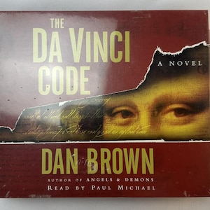 May include: A red, white, and black book cover with the title "The Da Vinci Code" and the author's name "Dan Brown". The cover features a close-up of a woman's eyes with a torn piece of paper revealing a handwritten note.