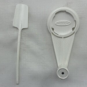 May include: Two white plastic kitchen utensils are shown against a textured background. One is a spatula with a rectangular head and a long handle. The other is a white bottle opener with the word "TOPPER" printed on it.