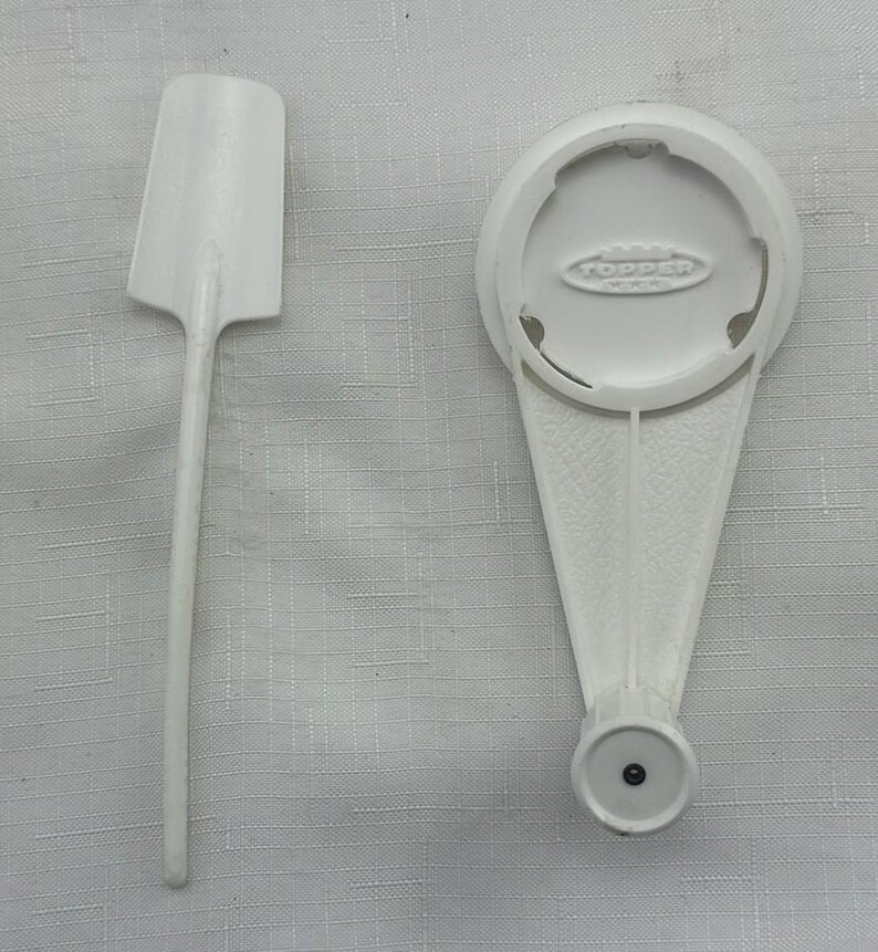 May include: Two white plastic kitchen utensils are shown against a textured background. One is a spatula with a rectangular head and a long handle. The other is a white bottle opener with the word "TOPPER" printed on it.
