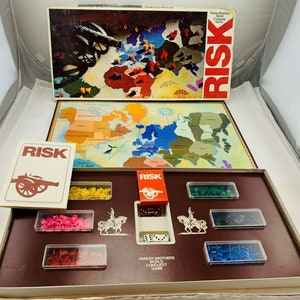 May include: A vintage RISK board game set. The box features a world map with a cannon illustration. The game includes a game board, cards, dice, and various colored game pieces. The game is made by Parker Brothers.