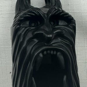 May include: A black, stylized face with pointed ears and a wide open mouth. The face has detailed features, including eyes, a nose, and visible teeth. The object is likely a decorative piece or a figurine.