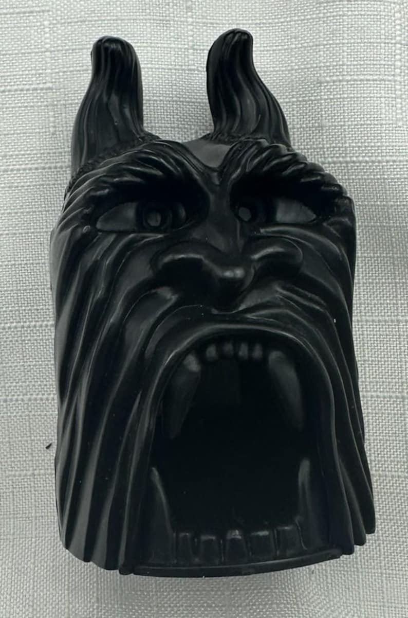May include: A black, stylized face with pointed ears and a wide open mouth. The face has detailed features, including eyes, a nose, and visible teeth. The object is likely a decorative piece or a figurine.