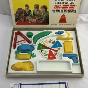 May include: Vintage board game titled "Pull the Rug Out Game." The box features the game's title and illustrations of children playing. Inside, various colorful game pieces are arranged, including a spinner, a foot-shaped piece, and a blue toy gun.
