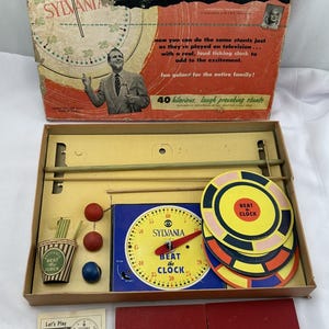 May include: A vintage board game called "Beat the Clock" by Sylvania. The game is based on the popular CBS television show. The box features a red, white, and blue color scheme with a clock face and a picture of the game show host. The game includes a spinning clock, a set of red and blue balls, and a set of yellow and black target discs.