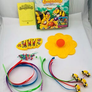 May include: A board game called "Bizzy Bizzy Bumblebees" with a yellow flower-shaped game board, bee-shaped game pieces, and a yellow instruction booklet. The game is a "Bee-Boppin' Pollen Poppin' Race Game".