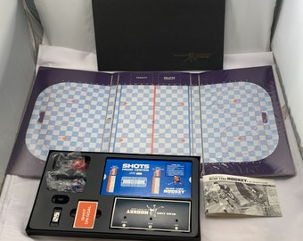 May include: A vintage Blue Line Hockey board game set. The set includes a game board with a hockey rink design, game pieces, dice, and a rule book. The game board is open, revealing the playing surface with blue and white checkered patterns.
