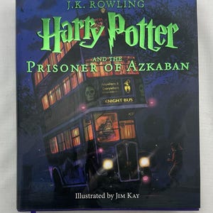 May include: Hardcover book titled "Harry Potter and the Prisoner of Azkaban" by J.K. Rowling, illustrated by Jim Kay. The cover features a purple Knight Bus with glowing windows and a running figure. The title is in green.