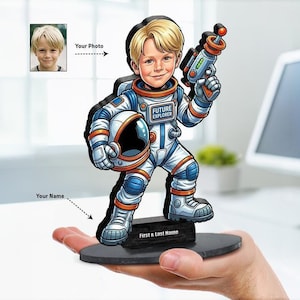 May include: A custom-printed wooden cutout of a cartoon astronaut with blonde hair, wearing a white and blue space suit with orange accents, holding a space helmet and a ray gun. The chest patch reads FUTURE EXPLORER and the base reads First & Last Name.