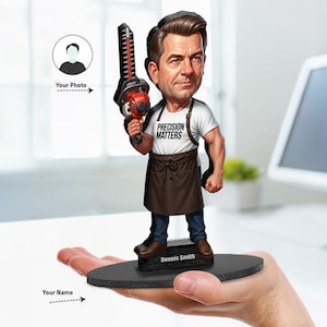 Op de afbeelding: Een gepersonaliseerde bobblehead-figuur met bruin haar, gekleed in een wit t-shirt met de tekst PRECISION MATTERS, een bruin schort en een blauwe spijkerbroek. Het figuur houdt een rood-zwarte heggenschaar vast en staat op een zwarte voet met de naam Dennis Smith.