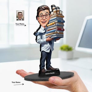May include: A custom caricature figurine of a person with glasses wearing a white shirt and dark trousers, holding a large stack of books and coffee cups. The figure stands on a black oval base with the name James Smith printed in white text.