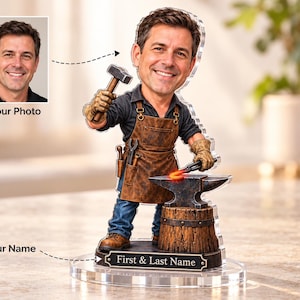 Könnte beinhalten: Eine personalisierte Wackelkopf-Figur eines Schmieds mit brauner Lederschürze, blauer Jeans und beigen Handschuhen. Die Figur hält einen Hammer und eine Zange über einem Amboss auf einem Holzstumpfsockel mit einem Schild First & Last Name.
