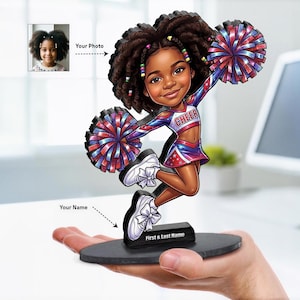 Puede incluir: Una figura de mesa personalizada de una animadora con cabello rizado oscuro, vistiendo un uniforme azul, rojo y blanco con la palabra CHEER en el pecho. La figura sostiene dos pompones y se apoya en una base negra con First & Last Name.