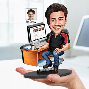 May include: A custom caricature figurine featuring a smiling person with dark hair sitting at a wooden desk with a computer, keyboard, and mouse. The figure wears a black and red shirt with blue jeans and stands on a black base labeled David Jackson.