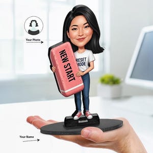 May include: A custom bobblehead figurine featuring a person with dark hair wearing a white t-shirt with the text RESET MODE and blue jeans. The figure holds a large pink eraser with the text NEW START and stands on a black base labeled Hannah.