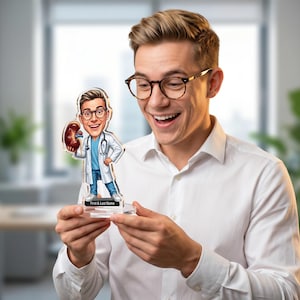 Op de afbeelding: Een gepersonaliseerd acrylbeeldje van een dokter met een nier. De cartoonarts draagt een witte jas, blauwe scrubs en een bril. De basis van het beeldje heeft ruimte voor aangepaste tekst. Het beeldje wordt vastgehouden door een persoon die een wit overhemd draagt.