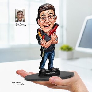 Może przedstawiać: Personalizowana figurka z kiwającą głową przedstawiająca osobę w okularach, czarnej koszuli i niebieskich dżinsach, trzymającą czerwoną suszarkę do włosów i przybory do stylizacji. Figurka stoi na czarnej owalnej podstawie z napisem James Smith.