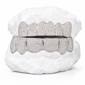 Może przedstawiać: Zestaw grillz wysadzanych diamentami, w srebrnym odcieniu, zakrywających górne i dolne zęby. Każdy ząb pokryty jest małymi, błyszczącymi diamentami. Grillz są prezentowane na białej, teksturowanej powierzchni.