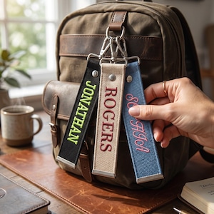 May include: Three rectangular fabric luggage tags attached to a brown backpack with silver metal clasps. The black tag features the name JONATHAN in lime green, the beige tag features ROGERS in dark red, and the blue tag features cursive text.