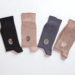 May include: Four pairs of socks in black, tan, and blue-grey, each with a unique embroidered monogram. The socks have a textured pattern and are displayed on a white surface. The sock cuffs and toes are in contrasting colors.